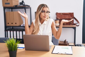 Young blonde woman working at the office wearing glasses clueless and confused expression with arms...