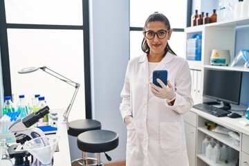 Young hispanic woman wearing scientist uniform using smartphone at laboratory