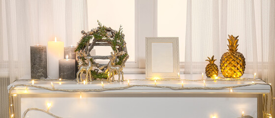 Christmas wreath with glowing lights and candles on mantelpiece near window in room