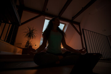 Half Lotus pose in dark lit room, mindfulness. Photo of young concentrated woman doing yoga exercises using tablet while sitting on floor at home.