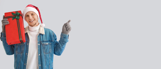 Handsome man in Santa hat holding Christmas present and pointing at something on light background with space for text