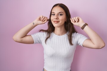 Young hispanic girl standing over pink background smiling pulling ears with fingers, funny gesture....