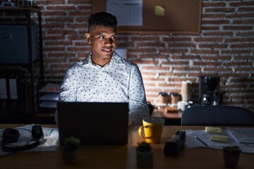 Young hispanic man working at the office at night looking away to side with smile on face, natural expression. laughing confident.