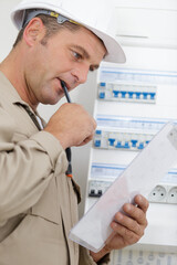 electrician stood by fusebox reading clipboard and chewing pen
