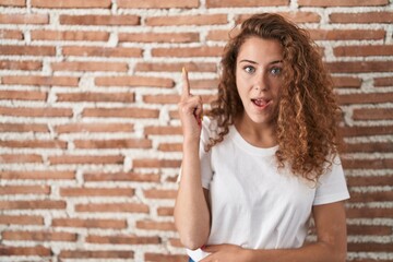 Young caucasian woman standing over bricks wall background pointing finger up with successful idea. exited and happy. number one.