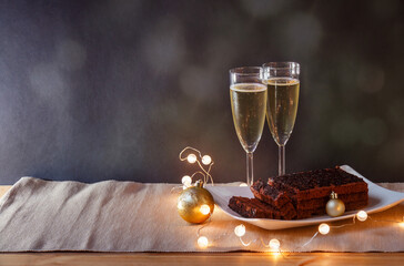 Two glasses of champagne, chocolate cake, glowing lights  and christmas decorations on table. Selective focus. View with copy space.