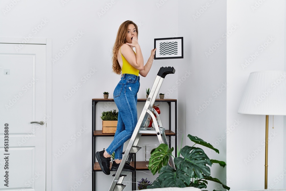 Poster Young caucasian woman on stairs hanging picture on the wall covering mouth with hand, shocked and afraid for mistake. surprised expression