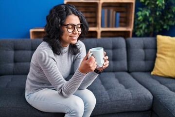 Young latin woman drinking coffee sitting on sofa at home