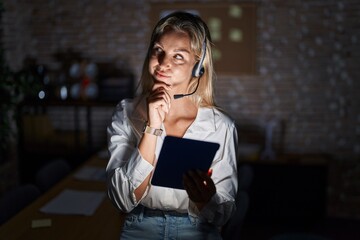 Young blonde woman working at the office at night with hand on chin thinking about question, pensive expression. smiling and thoughtful face. doubt concept.