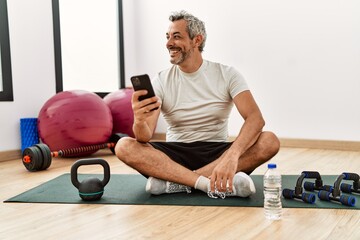 Middle age grey-haired man sitting on yoga mat using smartphone at sport center