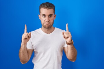 Young caucasian man standing over blue background pointing up looking sad and upset, indicating...