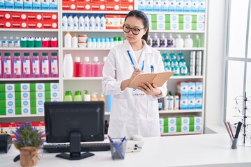 Young chinese woman pharmacist writing on notebook using computer at pharmacy
