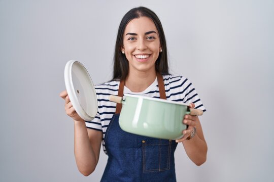 Young brunette woman wearing apron holding cooking pot smiling with a happy and cool smile on face. showing teeth.