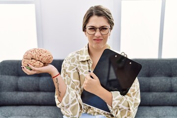 Young hispanic woman holding brain working at psychology clinic clueless and confused expression....