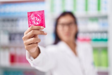 Young beautiful hispanic woman pharmacist smiling confident holding condom at pharmacy - obrazy, fototapety, plakaty