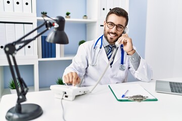 Young hispanic man wearing doctor uniform talking on the telephone at clinic