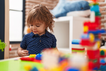 Adorable hispanic toddler playing with construction blocks standing at kindergarten