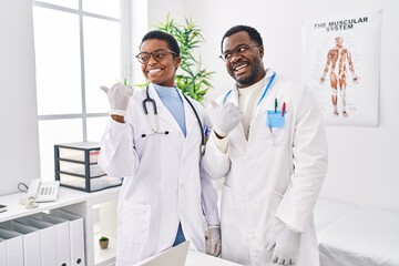 Young african american doctors working at medical clinic smiling with happy face looking and pointing to the side with thumb up.