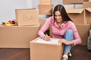 Young beautiful hispanic woman sitting on floor writing on paper at new home