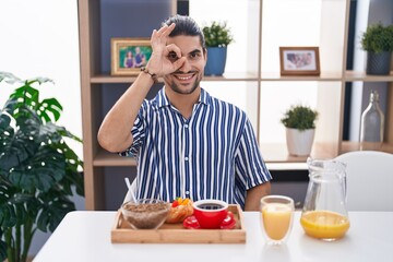 Hispanic man with long hair sitting on the table having breakfast smiling happy doing ok sign with hand on eye looking through fingers