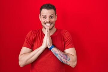 Young hispanic man standing over red background praying with hands together asking for forgiveness...