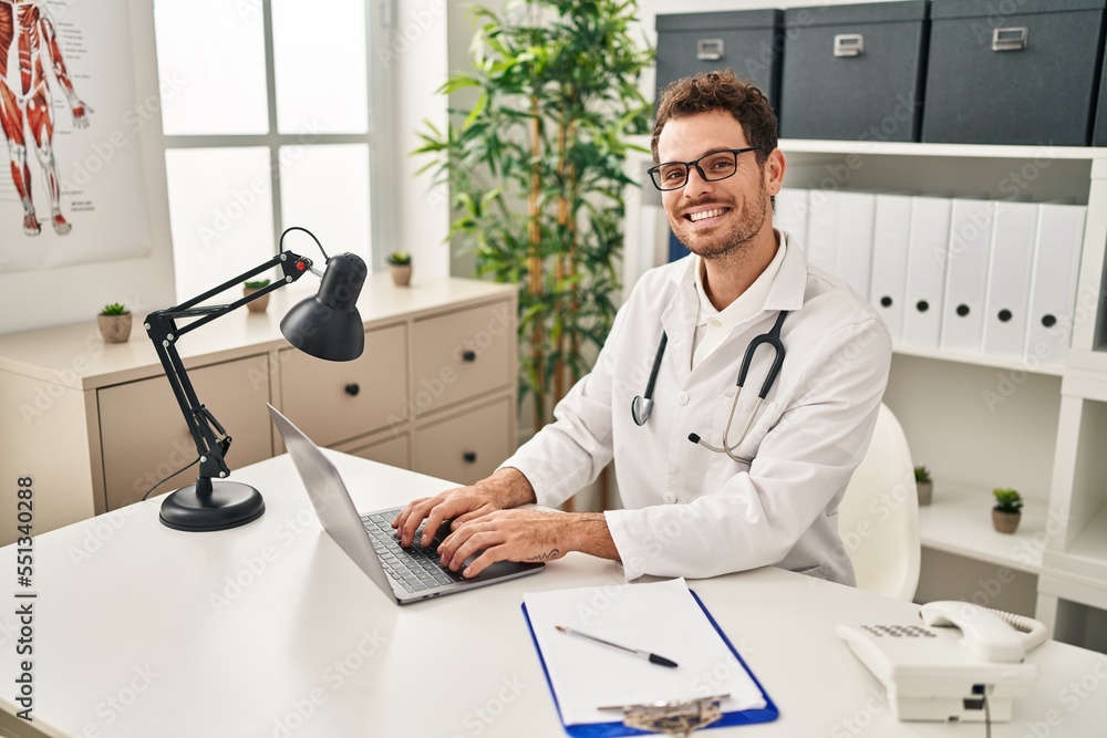 Canvas Prints Young hispanic man wearing doctor uniform using laptop at clinic