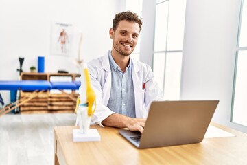 Young hispanic man wearing physiotherapist uniform using laptop working at clinic