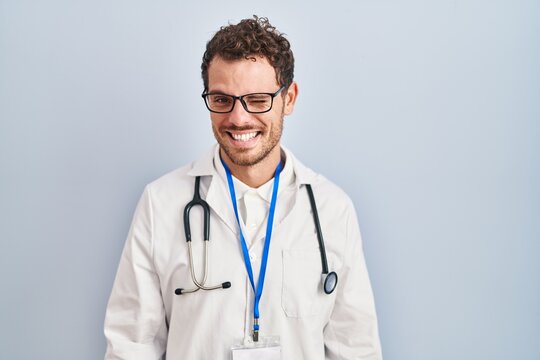 Young hispanic man wearing doctor uniform and stethoscope winking looking at the camera with sexy expression, cheerful and happy face.