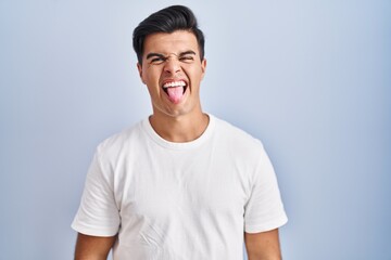 Hispanic man standing over blue background sticking tongue out happy with funny expression. emotion concept.