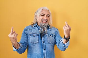 Middle age man with grey hair standing over yellow background showing middle finger doing fuck you...