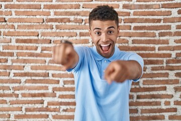 Brazilian young man standing over brick wall pointing to you and the camera with fingers, smiling positive and cheerful