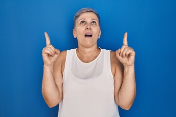 Middle age caucasian woman standing over blue background amazed and surprised looking up and pointing with fingers and raised arms.