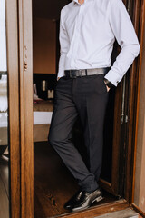 A stylish, rich, fashionable man, businessman, groom stands at the entrance near the wooden door in black pants, leather shoes and a white shirt. Photography, business.