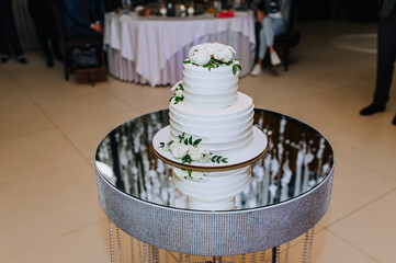 A beautiful white three-tier cake, decorated with flowers, roses, stands on a mirror table in a restaurant. Food photography, wedding.