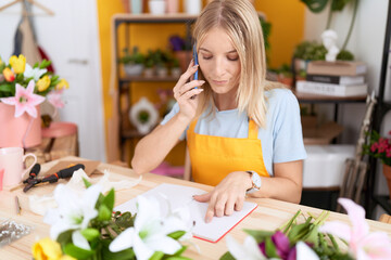 Young blonde woman florist talking on smartphone reading book at flower shop