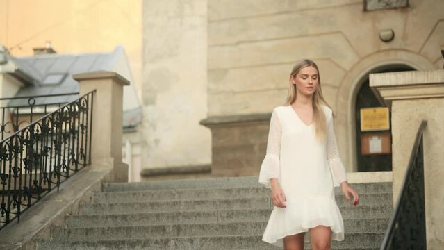 Beautiful young woman in a white dress is walking down the stairs in the city. Attractive fashion blonde model in a white dress with flying hair