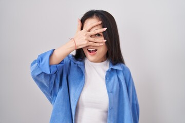 Young chinese woman standing over white background peeking in shock covering face and eyes with...