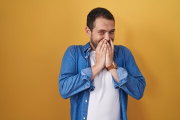 Hispanic man standing over yellow background laughing and embarrassed giggle covering mouth with hands, gossip and scandal concept