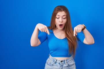 Fototapeta na wymiar Redhead woman standing over blue background pointing down with fingers showing advertisement, surprised face and open mouth