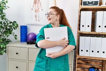 Young redhead woman wearing phsiologist uniform holding binder at clinic