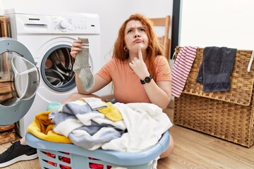Young redhead woman putting dirty laundry into washing machine thinking concentrated about doubt...