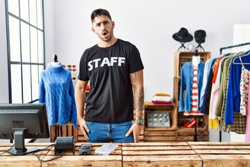 Young hispanic man working at retail boutique in shock face, looking skeptical and sarcastic,...