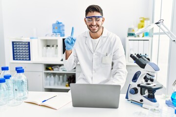 Young hispanic man working at scientist laboratory with laptop smiling with an idea or question pointing finger with happy face, number one