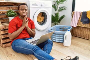 Young african man doing laundry and using computer begging and praying with hands together with hope expression on face very emotional and worried. begging.
