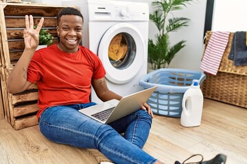 Young african man doing laundry and using computer showing and pointing up with fingers number three while smiling confident and happy.