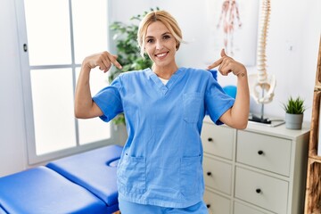 Beautiful blonde physiotherapist woman working at pain recovery clinic looking confident with smile on face, pointing oneself with fingers proud and happy.