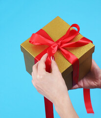 Female hands unpacking golden kraft paper gift box with red ribbon bow on isolated blue background