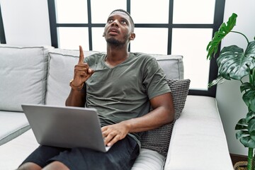 Young african american man using laptop at home sitting on the sofa amazed and surprised looking up and pointing with fingers and raised arms.