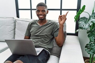 Young african american man using laptop at home sitting on the sofa with a big smile on face, pointing with hand finger to the side looking at the camera.