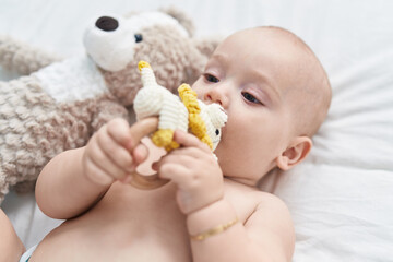 Adorable caucasian baby lying on bed biting doll at bedroom
